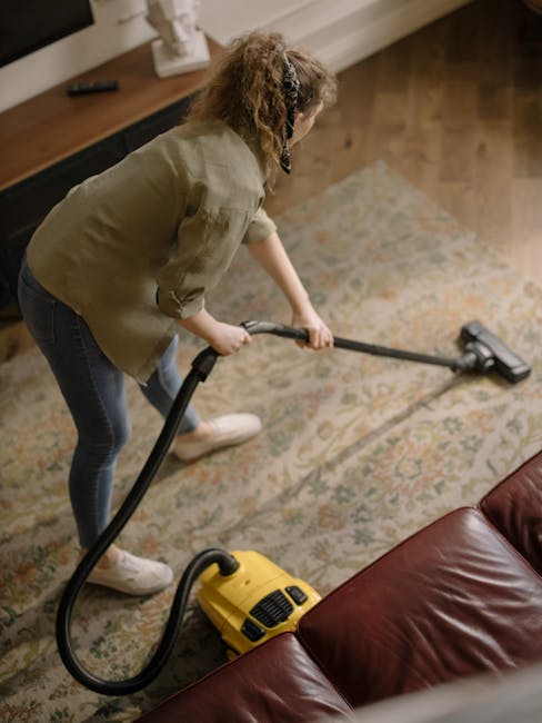 A woman in casual clothing, including a beige jacket and blue jeans, performing surface cleaning with a vacuum cleaner on a patterned area rug in a living room. The room features wooden flooring parti