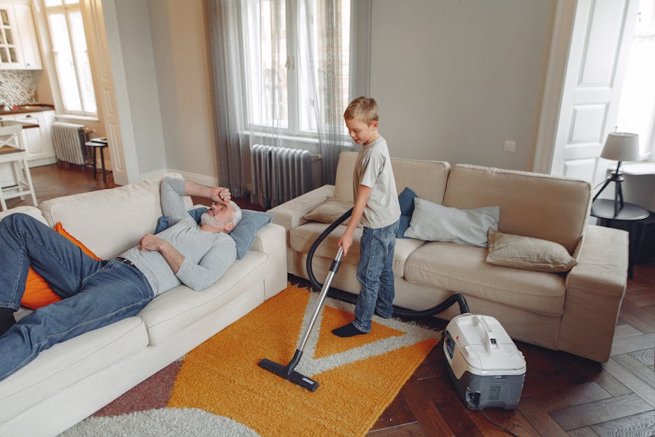 A woman in casual clothing, including a beige jacket and blue jeans, performing surface cleaning with a vacuum cleaner on a patterned area rug in a living room. The room features wooden flooring parti