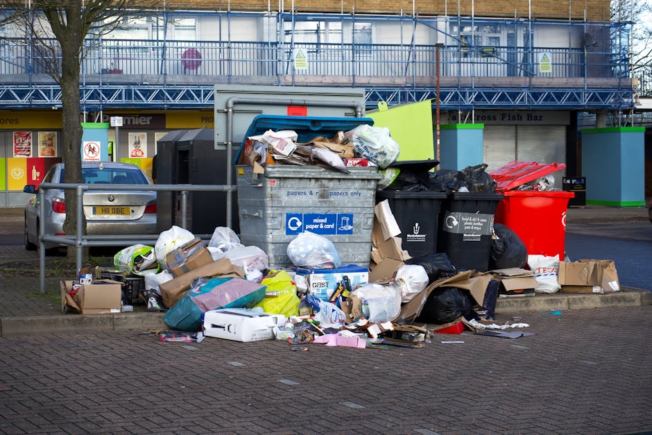 An outdoor scene showing overflowing waste bins and scattered rubbish bags on a sidewalk in front of a commercial building. The large grey bin labeled 'mixed paper & card' is open with cardboard boxes